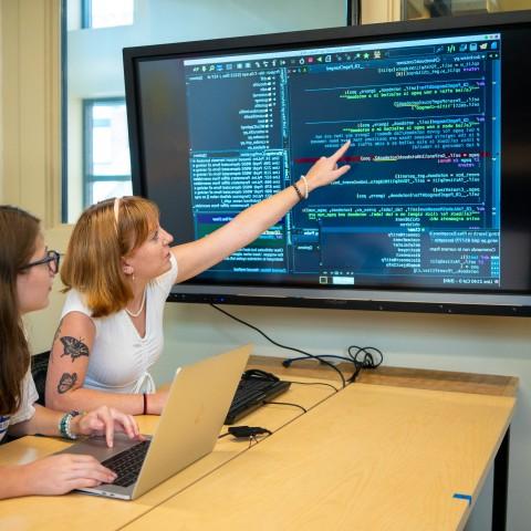 两个女学生坐在电脑前，电视屏幕上显示着大量的数据